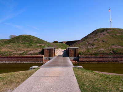 [Inbound view of wooden drawbridge over an approximately 20 foot wide moat. Paved walkway on either side of moat. Fort is out of view in this image.]